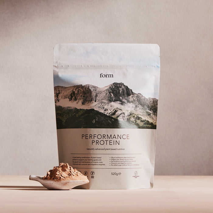 A Performance Protein bag by Form Nutrition is shown on a neutral backdrop with a mountain range image and text. This vegan, plant-based powder is perfect for post-workout recovery. A wooden scoop filled with the protein powder sits in front.