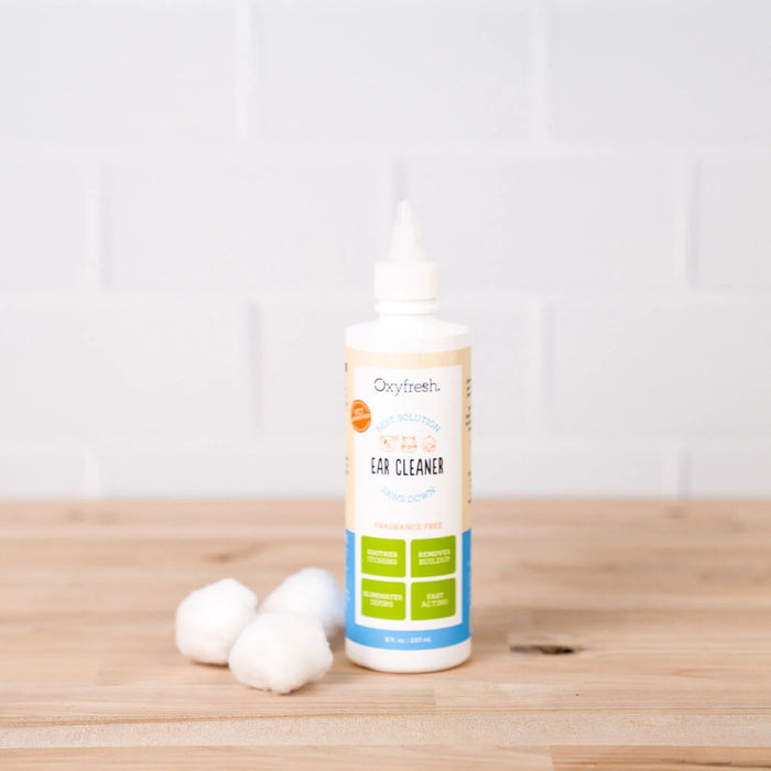 An Oxyfresh Pet Ear Cleaner Yeast-Fighting Ear Wash bottle sits on a wooden surface, accompanied by three cotton balls for your dogs ear cleaning routine against a backdrop of white brick.