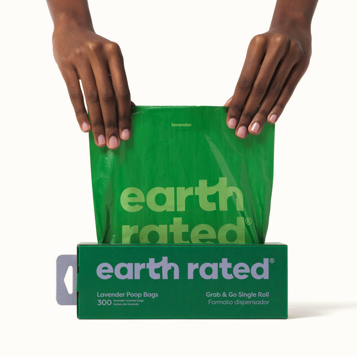 Hands pull a green bag from an Earth Rated box, showcasing lavender-scented, leak-proof pet waste bags. The packaging includes 300 large single roll refill bags and emphasizes its recycled plastic material against a white background.