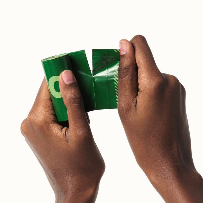Two hands pull apart a green Earth Rated pet waste bag wrapper featuring white and yellow lettering. Made from recycled plastic, the wrapper is torn in the middle against a plain white background.