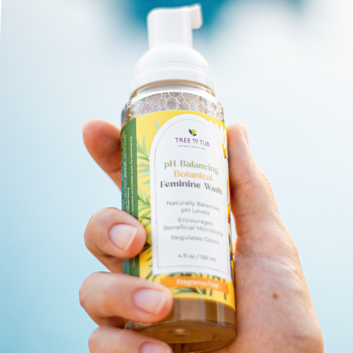 A hand holds a clear bottle of Tree To Tubs pH Balancing Botanical Feminine Wash, renowned for odor control and pH regulation. The labels benefits are highlighted against a soft, blurred light blue backdrop.