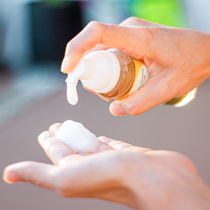 Close-up of a person dispensing Tree To Tubs pH Balancing Botanical Feminine Wash, a foamy liquid soap, from a bottle into their cupped hand. The blurred background emphasizes the focus on the hand and wash.