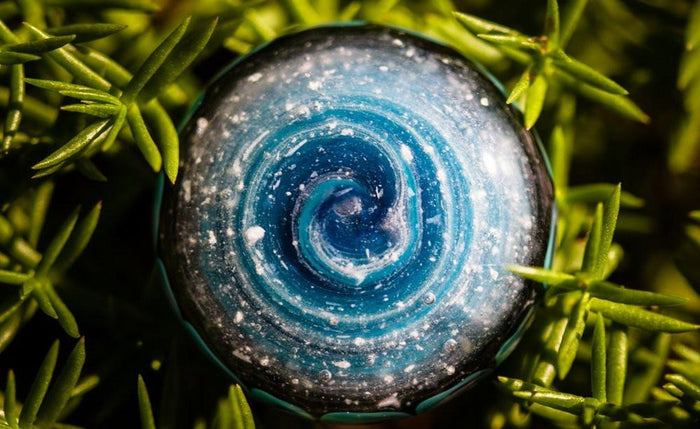 Top view of Aqua Pinwheel Touchstone infused with cremation ashes, blue swirls and speckled surface among green foliage.