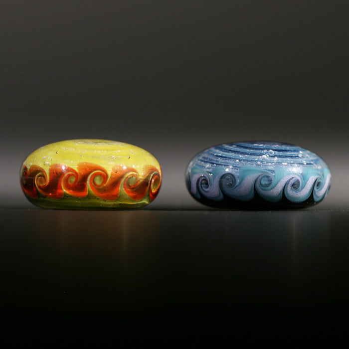 Side-by-side Pinwheel Touchstones on a dark surface, yellow with red swirls on the left and blue with teal swirls on the right.