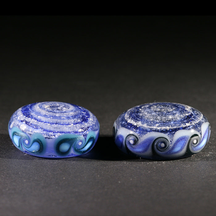 Two round pinwheel ash keepsakes infused with cremation ash in blue and white swirls on a dark surface