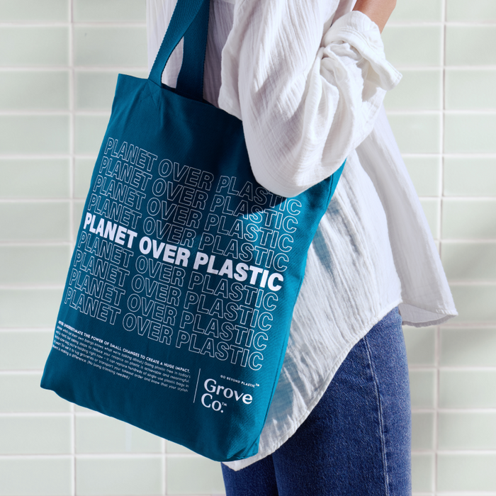 A person holds a Grove Co. Planet Over Plastic Reusable Tote Bag, its blue fabric adorned with white text, perfectly matching their white shirt and jeans against white tiled walls, highlighting their dedication to a plastic-free lifestyle.