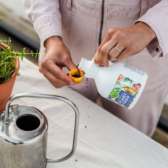 A person wearing a light jacket pours Good Dirts sustainable, chemical-free Plant Food into a cap. On the table sits a watering can and a terracotta pot with green herbs, as they ensure their plants flourish with this nourishment.