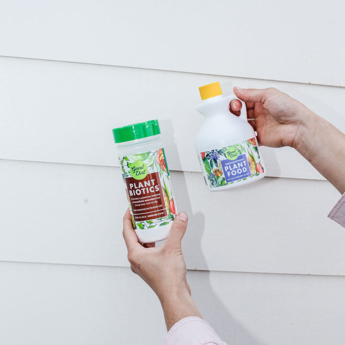 A person holds two gardening products against a white, paneled background: a bottle of chemical-free plant biotics with a green cap and Good Dirt Plant Food with a yellow cap.