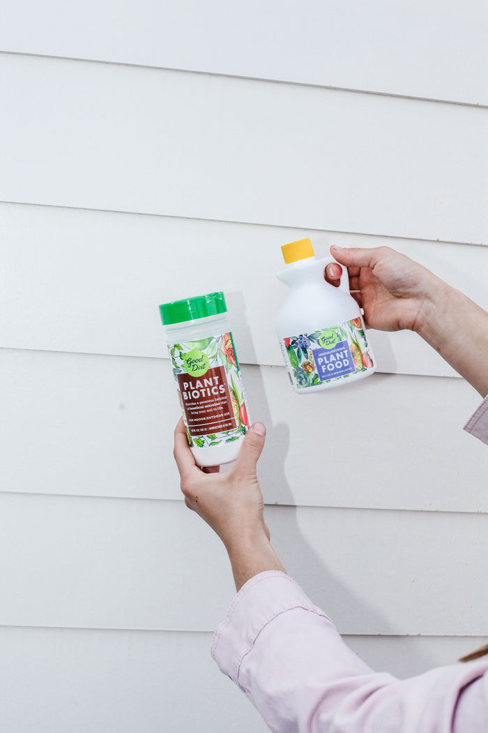 Two hands display garden products against a white wall. The left hand holds a container labeled Good Dirt PlantBiotic Indoor/Outdoor Nutrient Boost, promoting root development, while the right grips a bottle marked Plant Food to enhance nutrient uptake, with colorful plant illustrations on both.