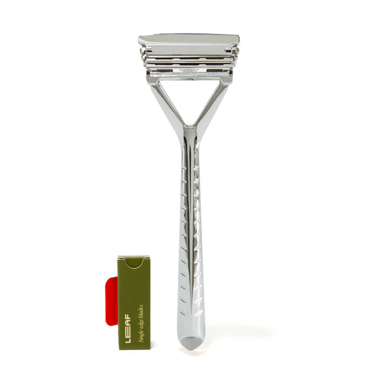 The Leaf Shave Plastic Free Razor is a sleek, polished safety razor with all-metal construction and a textured handle, ideal for sensitive skin. It stands upright next to a green and red box holding replacement blades.