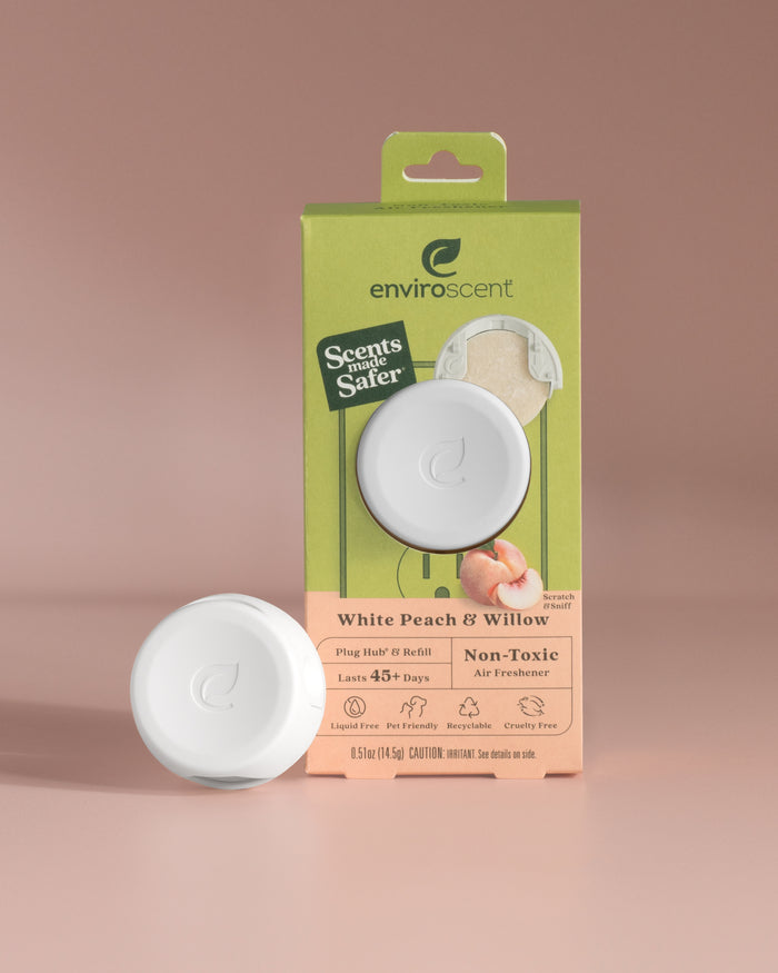 A white circular air freshener from the Enviroscent Plug-In Air Freshener Kit - Grove Exclusive stands against a soft pink background in front of its packaging that reads Enviroscent White Peach & Willow, promising a non-toxic, cruelty-free scent lasting over 45 days.