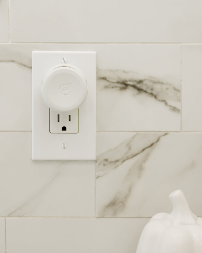 The Plug-In Air Freshener Kit - Grove Exclusive by Enviroscent is plugged into a wall outlet on a marble-tiled backsplash, with a white ceramic pumpkin peeking from the bottom right corner, creating a minimalist and clean setup.