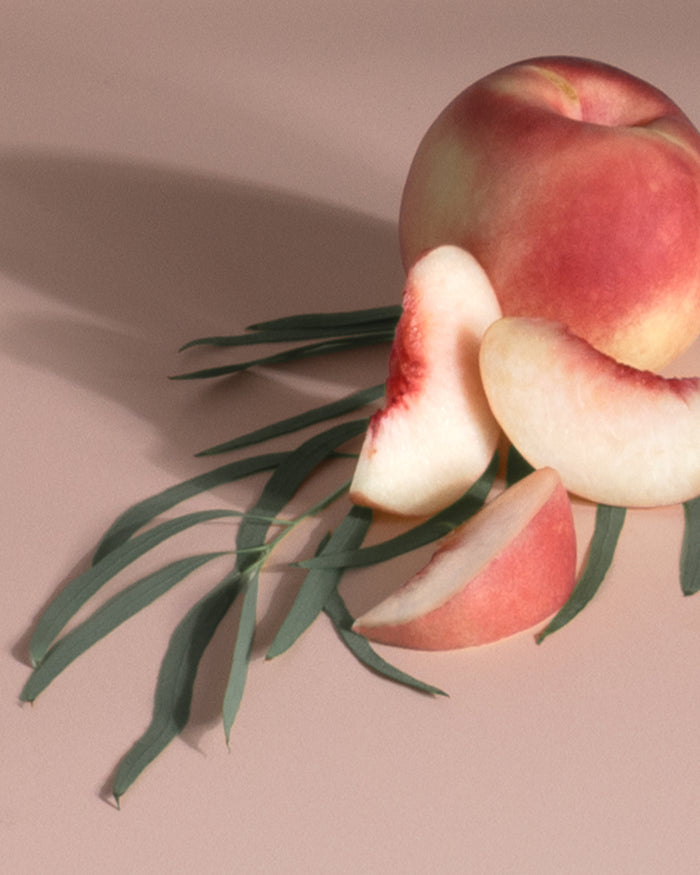 A blush-tinted peach rests on a peach surface with green leaves. Two slices reveal their soft, creamy interior, reminiscent of the soothing scent from Enviroscents Plug-In Air Freshener Kit - Grove Exclusive. Shadows softly extend across the surface.