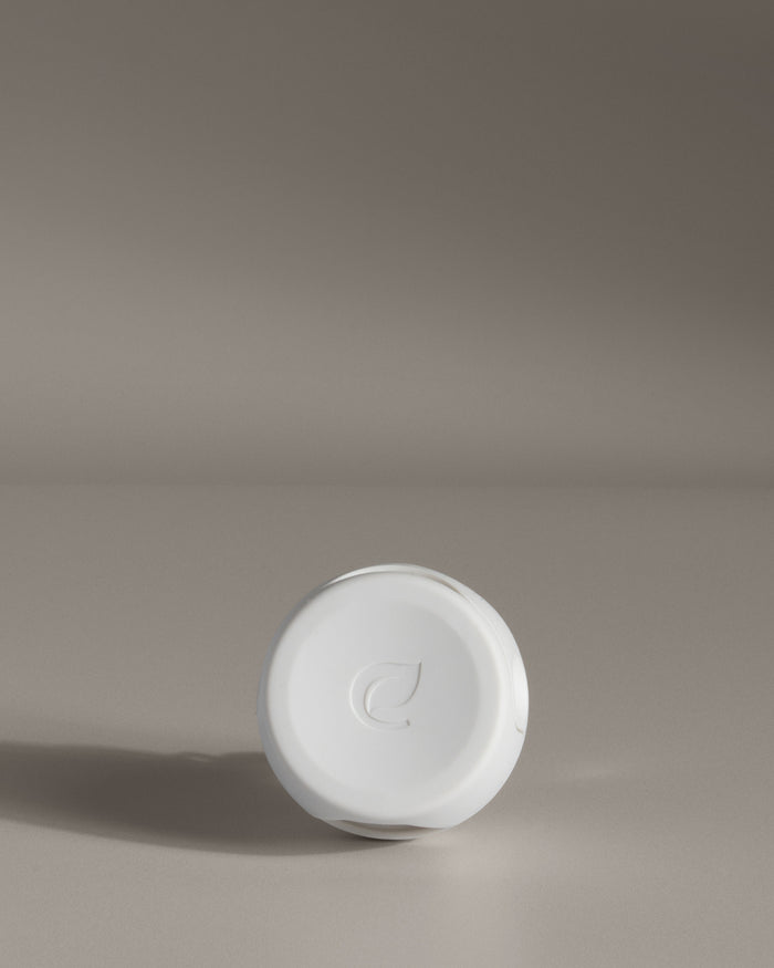 A white, round lid embossed with a leaf design from the Enviroscent Plug-In Air Freshener Kit - Grove Exclusive stands upright against a neutral background, perfect for sealing scent pod refills and casting a soft shadow.
