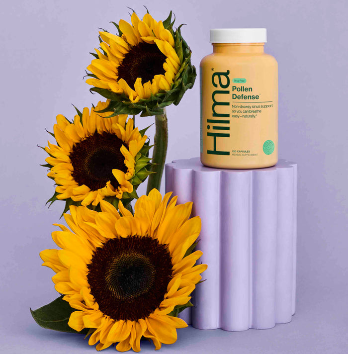 A Hilma Pollen Defense bottle, featuring natural herbs for sinus support, stands gracefully on a lavender pedestal beside three vibrant sunflowers against a calming light purple background.