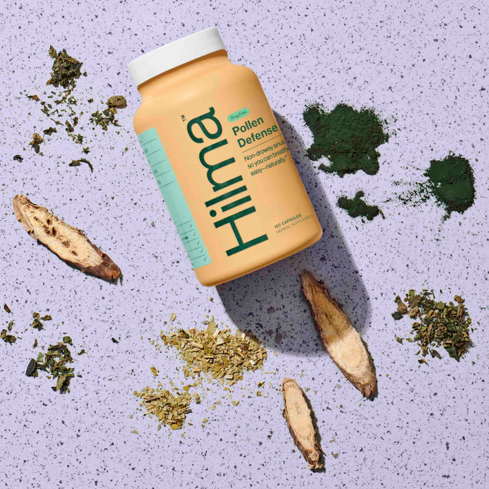 A bottle of Hilma Pollen Defense capsules rests on a speckled purple surface, surrounded by herbs, green powder, and plant roots, providing sinus support for those in need.