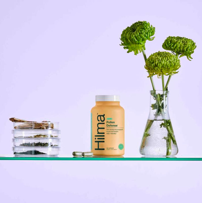 A Hilma Pollen Defense bottle is centered on a glass shelf. Green capsules for sinus support are on the left, with a small vase of green flowers to the right. Thin petri dishes containing dried natural herbs are farther left, all set against a light purple background.