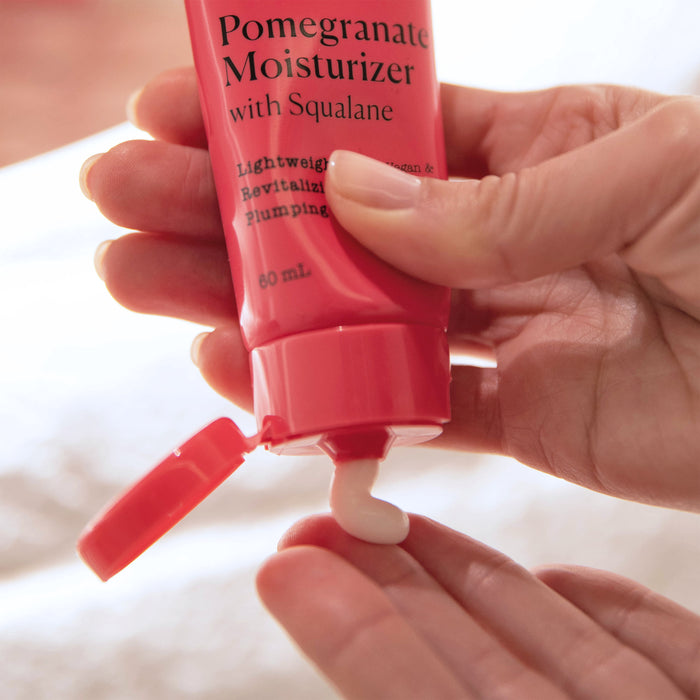 Hands squeeze a pink tube labeled APTO Skincare Pomegranate Moisturizer with Squalane, Lightweight Antioxidant Lotion onto fingers. This cruelty-free, hydrating cream is dispensed from an open cap, against a softly blurred background.
