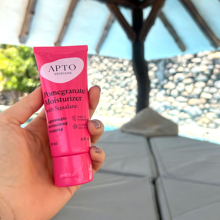 A hand holds a pink tube of APTO Skincares Pomegranate Moisturizer with Squalane, a lightweight antioxidant lotion thats cruelty-free. The outdoor backdrop features stone walls and a canopy, creating a soothing atmosphere.