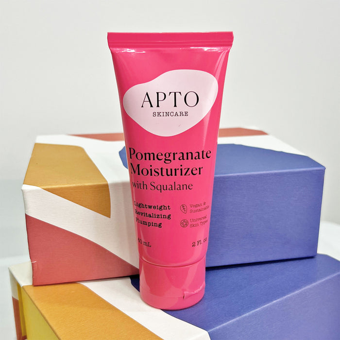 A pink tube of APTO Skincares Pomegranate Moisturizer with Squalane sits on vibrant geometric boxes, showcasing its cruelty-free, vegan packaging. This lightweight antioxidant lotion is unscented, plumping, and suitable for all skin types.