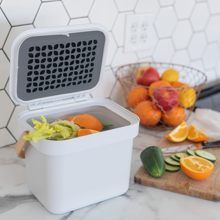 In a kitchen scene, the Full Circle Post Modern Compost Bin holds food scraps such as celery and oranges. A nearby fruit basket is brimming with citrus fruits, while a cutting board shows sliced cucumbers and oranges against a backsplash adorned with elegant hexagonal tiles.