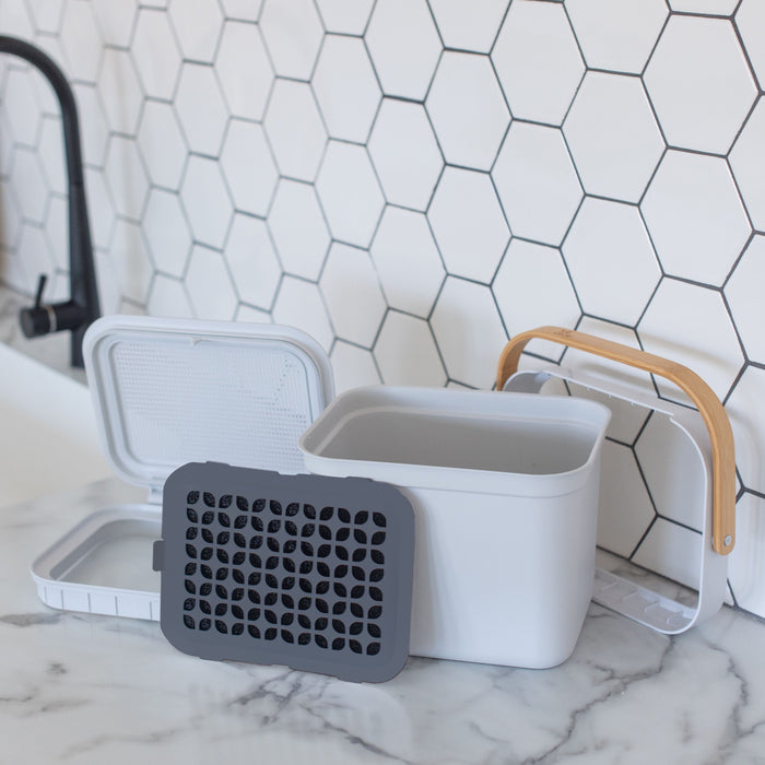 The Full Circle Post Modern Compost Bin with a wooden handle is on a marble countertop. The open lid reveals an activated carbon filter. A white hexagonal-tiled wall and sleek black faucet create a stylish background.