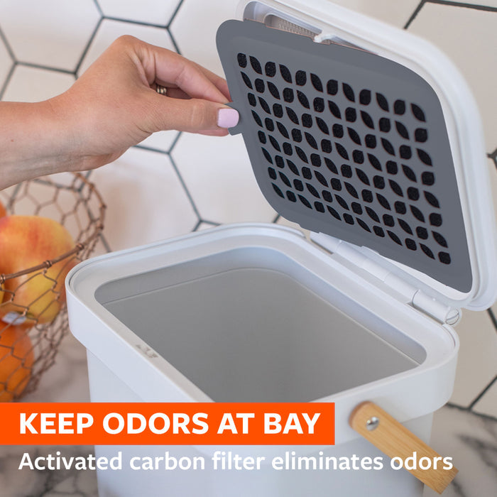 A person opens a small white compost bin with a gray activated carbon filter inside the lid. The bin is placed on a counter with an orange fruit nearby. An orange banner at the bottom reads, "KEEP ODORS AT BAY. Activated carbon filter eliminates odors.