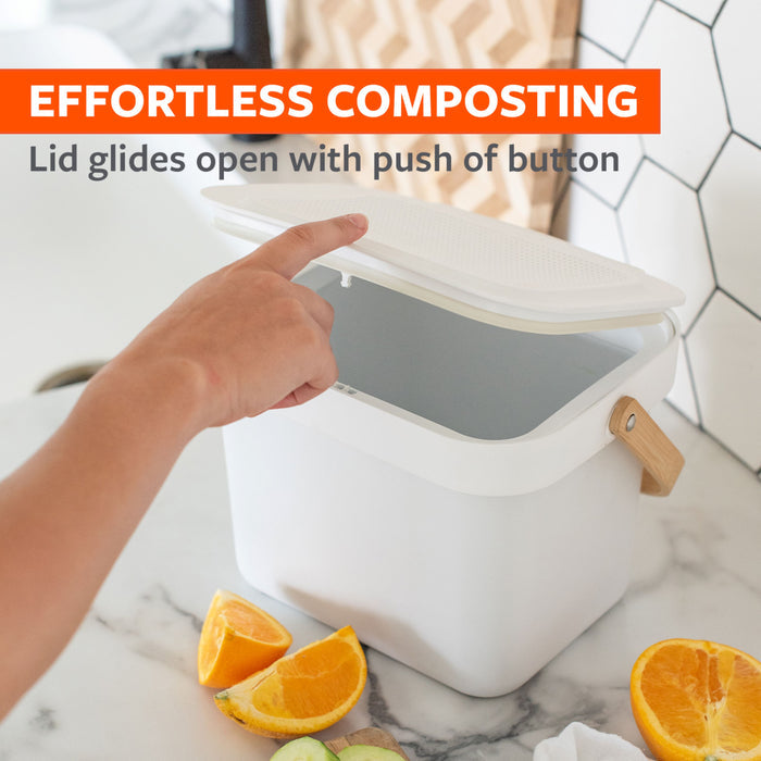 A hand opens the lid of a white compost bin on a kitchen counter. The text reads, "EFFORTLESS COMPOSTING: Lid glides open with push of button." Orange slices and peels are on the counter beside the bin.