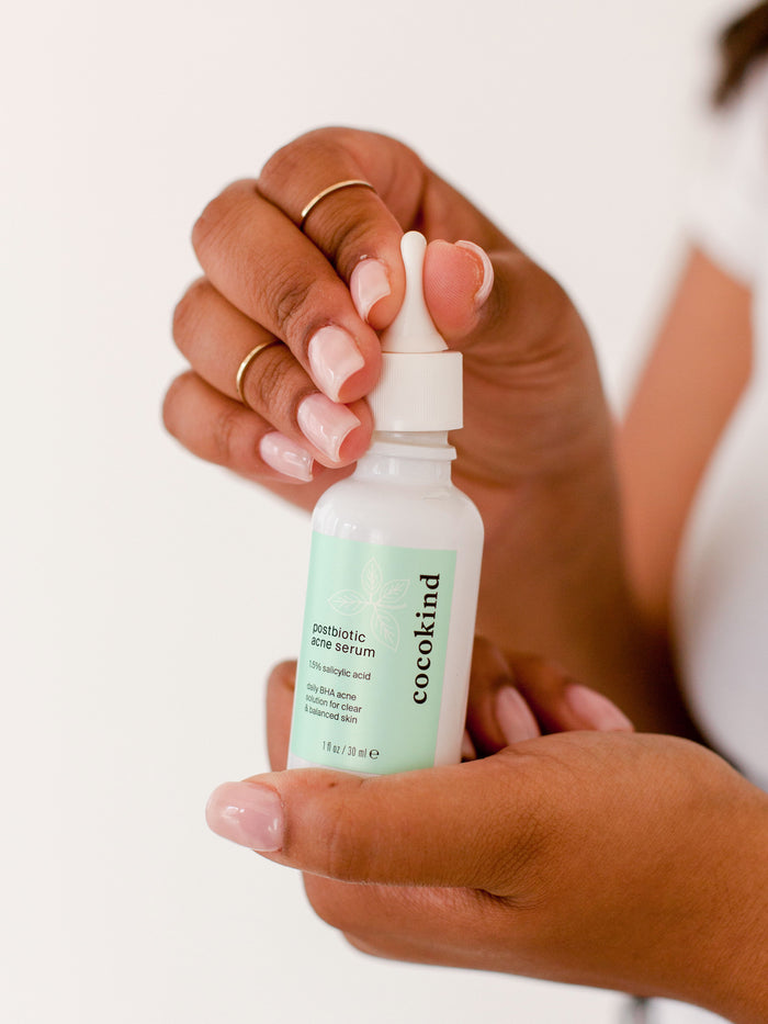 A person with manicured nails holds a small Cocokind Postbiotic Acne Serum bottle. Perfect for clogged pores, it has a dropper top and features a light blue label on a plain white background.