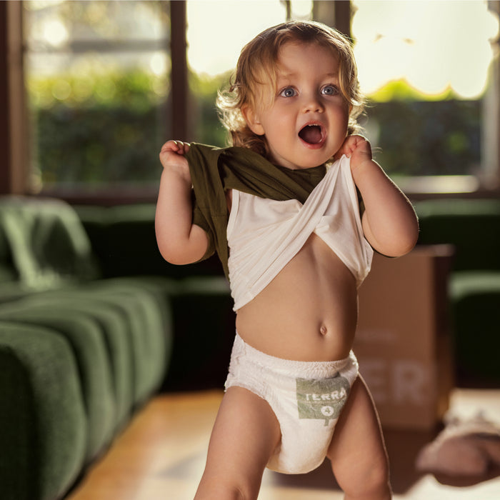 In a sunlit room, a toddler playfully attempts to put on Terras 100% bamboo fiber Potty Pull Up Training Pants. A green couch and a plant-based cardboard box add an eco-friendly touch to this charming scene.