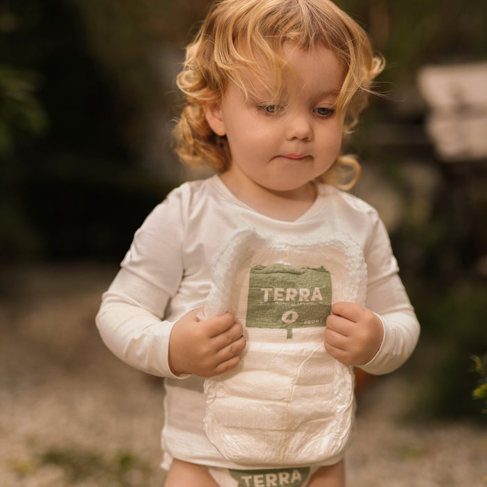 A toddler with curly blond hair, wearing a white shirt, stands outdoors holding Terra Potty Pull Up Training Pants against their chest, with greenery and a blurred stone surface in the background.