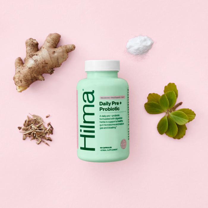 A bottle of Hilmas Pre + Probiotic sits on a pink backdrop with ginger root, white powder, green leaves, and shredded natural material around it—emphasizing its potent blend for improved gut health.