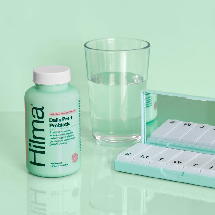 A green Hilma Pre + Probiotic bottle for gut support is next to a glass of water and a mint pill organizer marked with days, all on a reflective mint surface against a matching background.