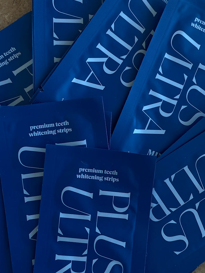 A collection of Plus Ultra Premium Teeth Whitening Strips in blue packets, ideal for sensitive teeth, is scattered randomly with partial overlap creating a layered effect.