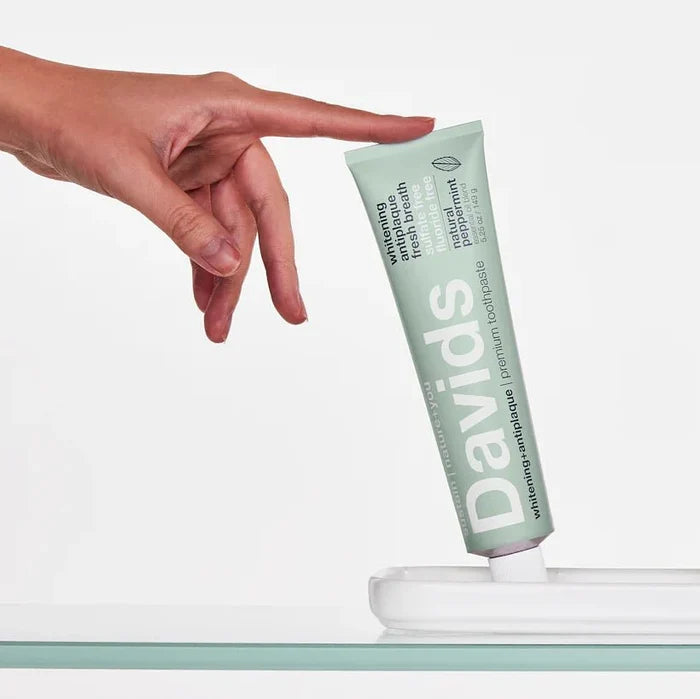 A hand reaches for a green tube of Davids Premium Toothpaste - Whitening + Antiplaque, which is standing upright on a white holder against a plain white background.