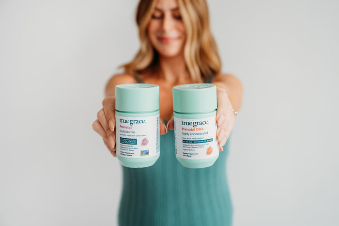 A woman with blurred features holds True Grace Prenatal DHA and Multivitamin supplements, boosting omega-3s for brain and eye health. Both feature a light blue-green lid and label. Dressed in teal, she stands against a plain backdrop.