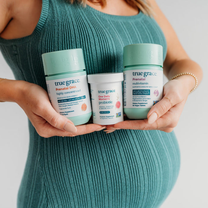 A person in a teal ribbed dress holds True Graces Prenatal DHA known for high-quality omega-3s, along with One Daily Womens Probiotic and Prenatal Multivitamin, all set against a plain white backdrop.