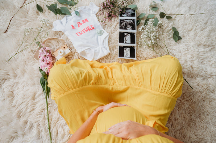 A person in a yellow dress sits on a fluffy rug, cradling their belly, nourished by True Graces Prenatal DHA. Nearby are a baby onesie with I AM FUTURE printed on it, a strip of ultrasound photos, flowers, and tiny baby shoes.