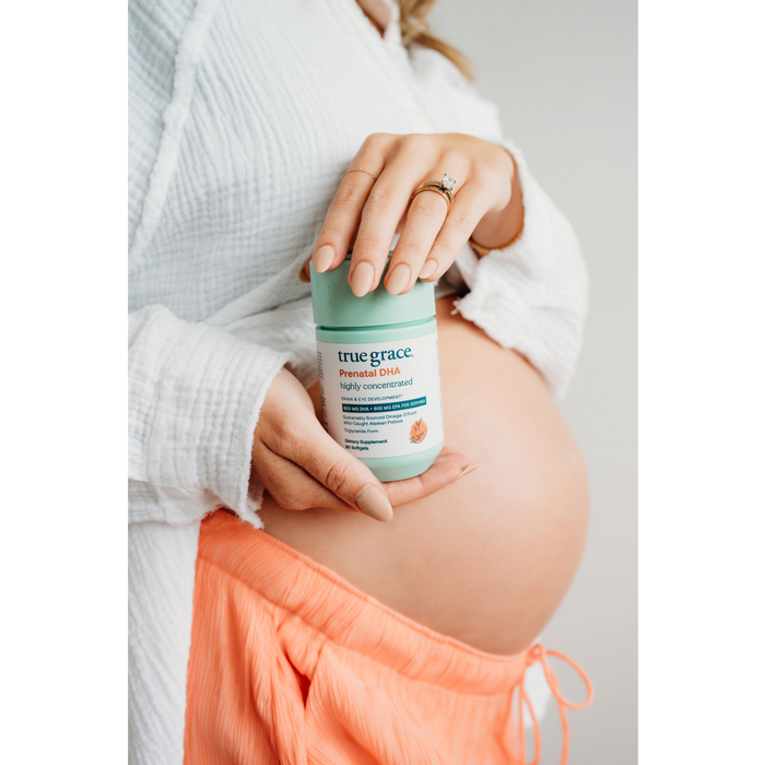 A pregnant individual in an unbuttoned white shirt and orange pants showcases True Graces Prenatal DHA, rich in Omega-3s, holding it against their bare belly while highlighting their engagement ring.