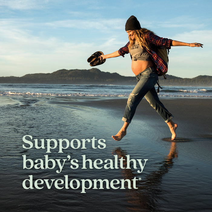 A pregnant woman joyfully leaps barefoot across a shallow stream at the beach. She wears jeans, a plaid shirt, a knit hat, and carries boots and a backpack. The sky is clear, with hills in the background. Text reads, "Supports baby's healthy development*".