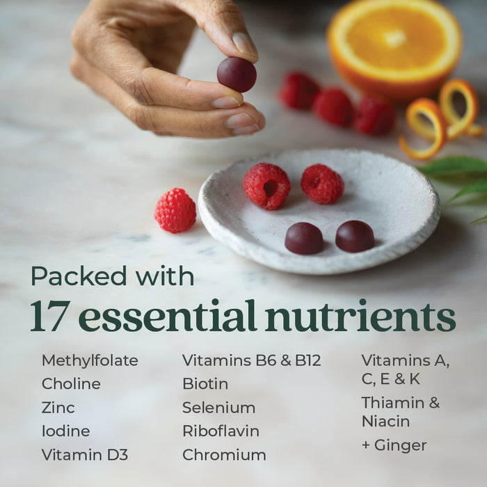 A hand places three red gummies on a small white plate. Beside them are raspberries and sliced oranges. Text reads: "Packed with 17 essential nutrients," followed by a list including vitamins, minerals, and ginger.