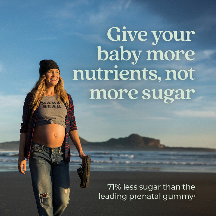 A pregnant woman walks on a beach holding sandals, wearing a "Mama Bear" shirt and plaid overshirt. Text reads: "Give your baby more nutrients, not more sugar. 71% less sugar than the leading prenatal gummy." A coastline is visible in the background.