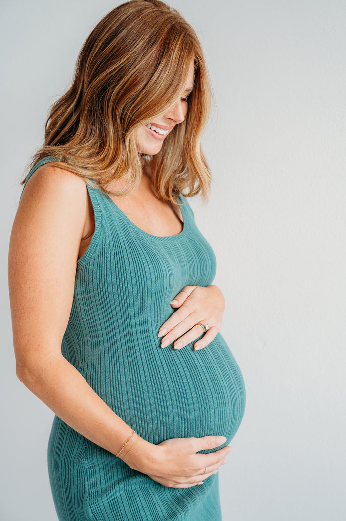 A pregnant woman with long brown hair gently smiles, looking down and cradling her belly. Wearing a teal sleeveless dress against a plain light background, she embodies the glow of expecting mothers who trust True Graces Prenatal Multivitamin.
