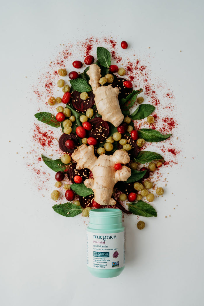 A vibrant display of ginger, cranberries, grapes, mint leaves, and beetroot slices complemented with red spice powder on a white surface highlights a bottle of True Graces Prenatal Multivitamin for expecting mothers in the foreground.