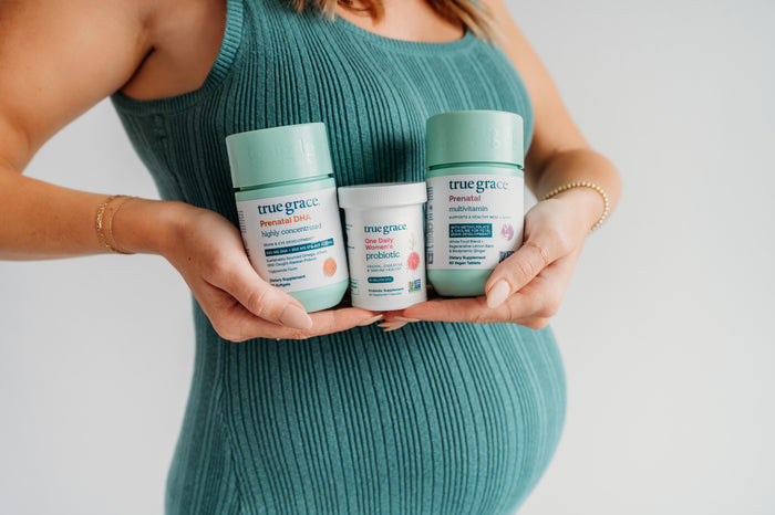 A pregnant woman in a teal ribbed dress holds three True Grace supplement containers: Prenatal DHA, One Daily Probiotic, and Prenatal Multivitamin with added Choline, against a plain light background.