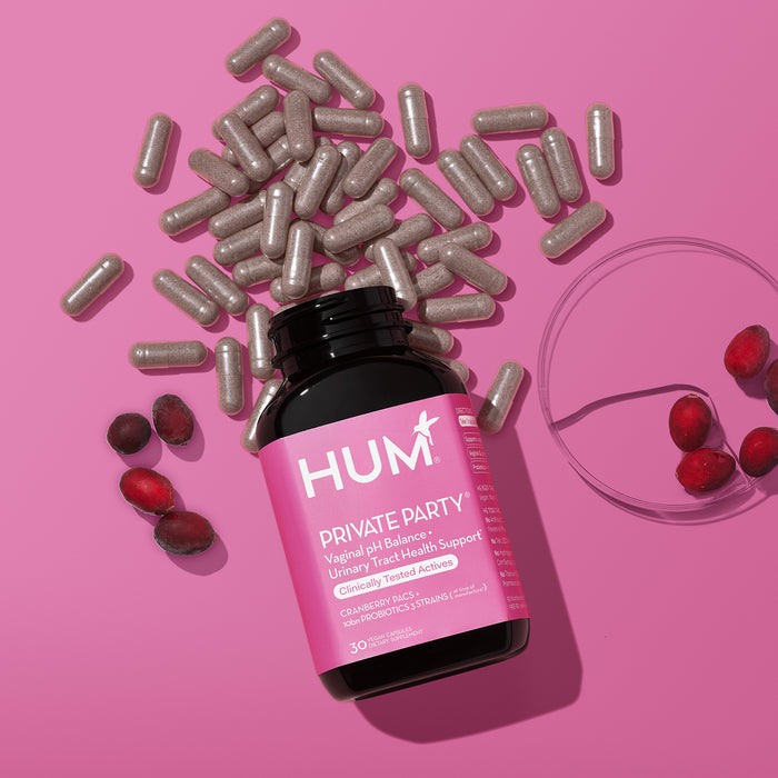 An open pink bottle of HUM Nutritions Private Party Probiotics for Vaginal Health sits on a pink background, surrounded by scattered capsules and whole cranberries. Nearby, a petri dish holds more cranberries, emphasizing their health benefits.