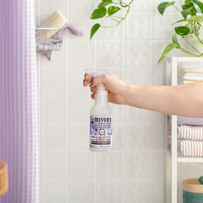 A hand holds a bottle of Mrs. Meyers Probiotic Daily Shower Spray Cleaner in a bathroom with lavender scent. The white tiled walls, potted plant, and neatly stacked towels in the background make it perfect for tackling soap scum stylishly.