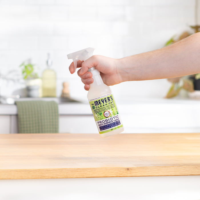 A hand holds a Mrs. Meyers Probiotic Multi-Surface Cleaning Spray over wood, with a blurred kitchen in the background. The invigorating Lemon Verbena scent enhances your cleaning routine.
