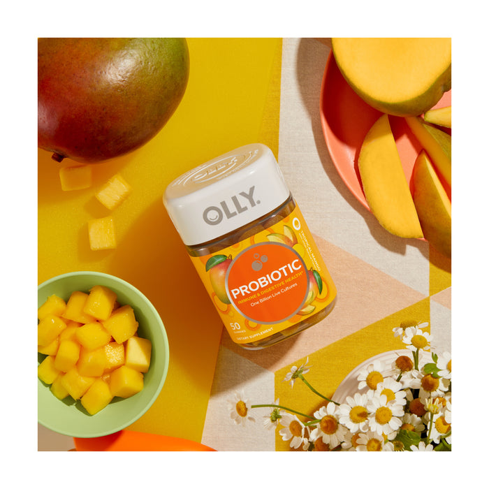 Top view of a vibrant display featuring a jar of OLLY Probiotic gummies for gut health, surrounded by fresh mango slices, chunks in a bowl, a whole mango, and small white flowers on a geometric-patterned background.