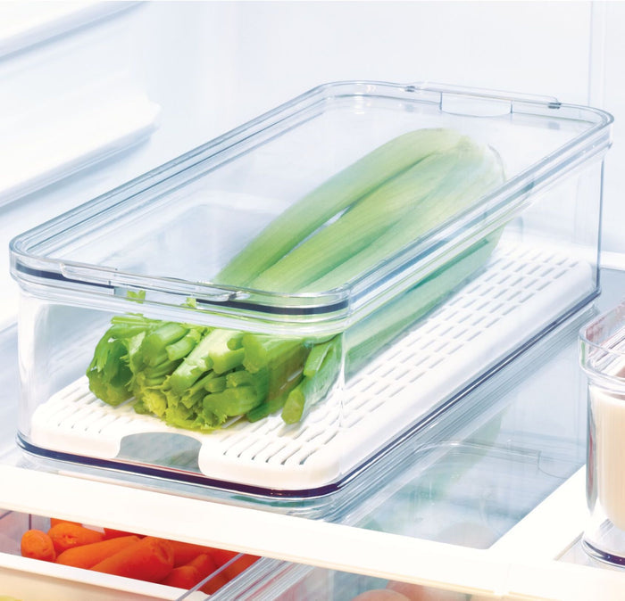 The iDesign Produce Fridge Bin, made with recycled BPA-free plastic, holds celery in the refrigerator with a transparent lid and white tray base. In the bottom left, another bin with carrots is partially visible.